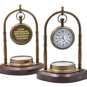 May include: Two antique brass pocket watch stands with a compass on the base. The pocket watch on the right has a white face with black Roman numeral hour markers and black hands. The pocket watch on the left has a gold face with black text that reads "2nd Annual PrepareRI Employer Awards Rookie of the Year Award Rhode Island Department of Business Regulation".