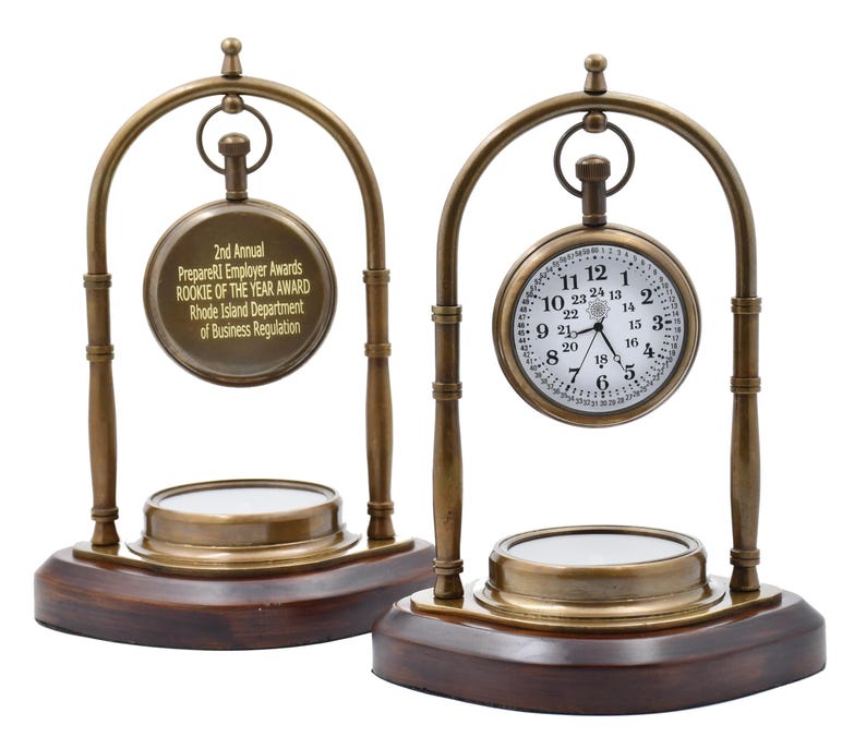 May include: Two antique brass pocket watch stands with a compass on the base. The pocket watch on the right has a white face with black Roman numeral hour markers and black hands. The pocket watch on the left has a gold face with black text that reads "2nd Annual PrepareRI Employer Awards Rookie of the Year Award Rhode Island Department of Business Regulation".