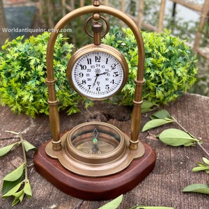 May include: A brass pocket watch with a compass on a wooden base. The watch face has black Roman numerals and a white background. The compass has a green needle and a white background.