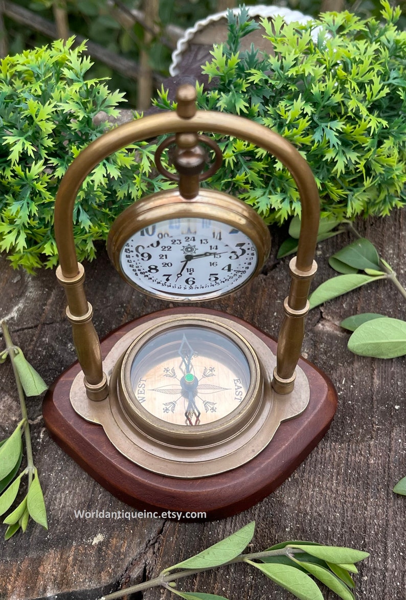 May include: A brass compass and pocket watch on a wooden stand. The compass has a green jewel in the centre and the watch has Roman numerals and Arabic numerals.