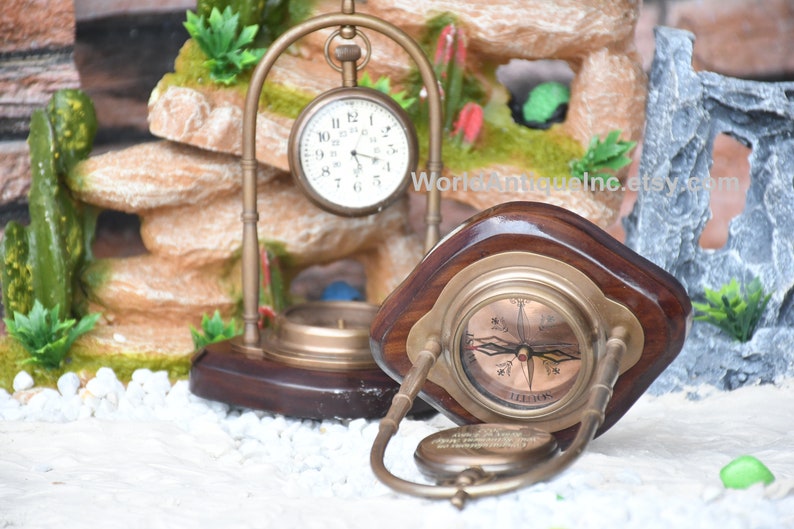 May include: A brass compass and pocket watch on wooden stands. The compass has a white face with black markings and a red needle. The pocket watch has a white face with black Roman numeral markings and black hands. The stands are made of dark wood with a brass trim.
