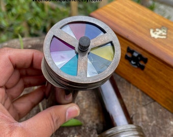 May include: A brass colored color wheel with a rotating disc of colored glass. The wheel is attached to a brass tube. The tube is sitting on a wooden surface. The text "Worldantiqueinc.etsy.com" is visible in the image.