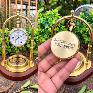 May include: A brass desk clock and compass set mounted on a dark wood base. The clock features a white face with black numerals and a 24-hour scale. The back of the clock is engraved with the text A Love That's Timeless II-XXIV-MMXXIV.
