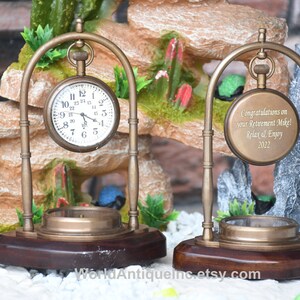 May include: Two antique brass pocket watch stands with a wooden base. The watch on the left has a white face with black Roman numeral hour markers. The watch on the right has a brass face with an engraved message that reads "Congratulations on your Retirement Mike! Relax & Enjoy 2022".