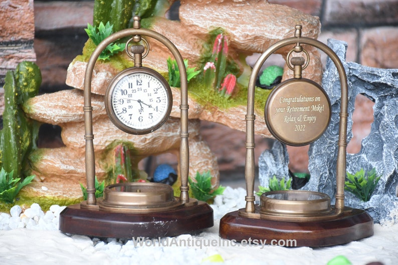 May include: Two antique brass pocket watch stands with a wooden base. The watch on the left has a white face with black Roman numeral hour markers. The watch on the right has a brass face with an engraved message that reads "Congratulations on your Retirement Mike! Relax & Enjoy 2022".
