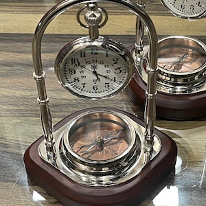 Pode incluir: Um suporte de metal prateado com uma base de madeira segura dois rel&oacute;gios de bolso e uma b&uacute;ssola. Os rel&oacute;gios de bolso t&ecirc;m mostradores brancos com marcadores de hora romanos pretos e ponteiros pretos. A b&uacute;ssola tem um mostrador de cor cobre com uma agulha preta.