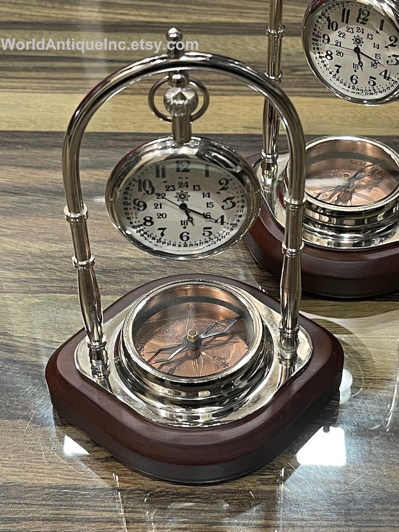 Pode incluir: Um suporte de metal prateado com uma base de madeira segura dois rel&oacute;gios de bolso e uma b&uacute;ssola. Os rel&oacute;gios de bolso t&ecirc;m mostradores brancos com marcadores de hora romanos pretos e ponteiros pretos. A b&uacute;ssola tem um mostrador de cor cobre com uma agulha preta.