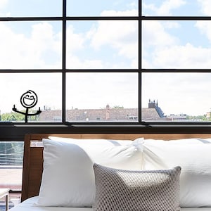 May include: A black and white stick figure with a smiling face and raised arms is positioned in front of a window. The window has a grid pattern and overlooks a cityscape with a cloudy sky. The scene includes a bed with white pillows and a decorative pillow.