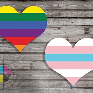 Puede incluir: Dos diseños en forma de corazón sobre un fondo de madera. El primer corazón es un arcoíris con rayas rojas, naranjas, amarillas, verdes, azules, índigo y violetas. El segundo corazón es una bandera del orgullo transgénero con rayas azul claro, rosa y blancas.