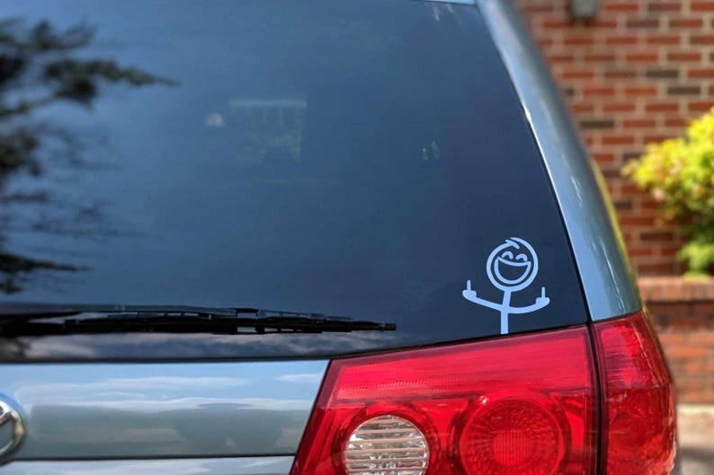 May include: A white vinyl car decal featuring a stick figure with a smiling face and raised arms, displaying the middle finger on each hand. The decal is affixed to the rear window of a silver vehicle.