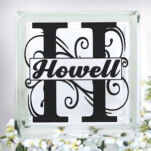 May include: A clear glass block with a black monogram letter "H" and the name "Howell" in a decorative font. The design includes swirling flourishes. The block is decorated with white flowers.