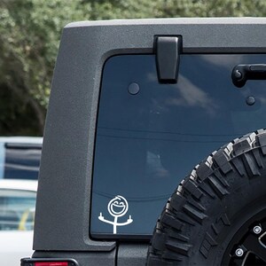 May include: White vinyl decal of a stick figure with a smiling face and raised middle fingers. The decal is applied to the rear window of a dark gray vehicle, with a black tire visible in the lower right corner.