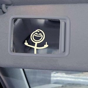 May include: A car sun visor with a built-in mirror. The mirror features a yellow stick figure with a smiling face and raised middle fingers. The visor is gray and the mirror is rectangular.