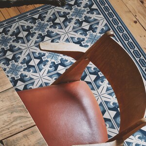 Peut inclure: Un tapis de sol bleu et blanc à motifs géométriques. Le tapis est placé sur un sol en bois et une chaise en bois est posée dessus. La chaise a un siège en cuir marron.