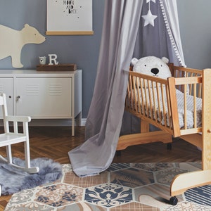 Könnte beinhalten: Ein Babybett aus Holz mit einem weißen Stofftier darin, einem grauen Baldachin und einem weißen Schaukelstuhl im Vordergrund. Das Bett steht auf einem gemusterten Teppich mit einem grauen und weißen Teppich darunter. Der Raum hat eine hellblaue Wand mit einer weißen Bärensilhouette an der Wand.