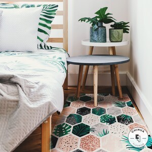 May include: A bedroom featuring a bed, two bedside tables, and a patterned rug. The bed has a white duvet and green and white patterned pillows. The bedside tables are made of wood and have white tops. The rug is a geometric pattern in pink, grey, and black. The rug has a logo that says "Chinua Flooring".