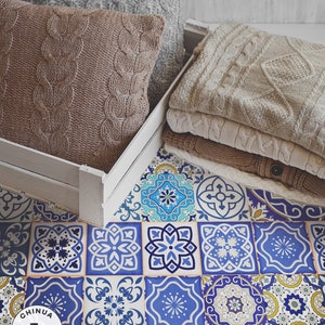 Puede incluir: Una caja de madera blanca con dos almohadas de punto marrón y una pila de jerséis de punto beige. La caja está sentada en un suelo de baldosas con estampado azul y blanco. El suelo tiene el texto "CHINUA FLOORING" en negro.