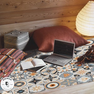 Puede incluir: Un espacio de vida acogedor con una cama que presenta un diseño de baldosas estampadas en tonos azules, naranjas y grises. Un portátil, libros y almohadas están dispuestos en la cama, creando un ambiente relajado. El texto "CHINUA FLOORING" es visible en la esquina inferior izquierda de la imagen.