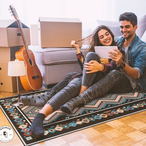 May include: A couple sits on a black and blue patterned rug with a geometric design. The woman is holding a tablet computer and the man is looking at the tablet. They are both smiling.