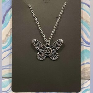 May include: A silver butterfly necklace with a silver triangle charm in the center. The charm has a circle around it with a symbol inside. The necklace is on a black background. "If nothing ever changed, there would be no butterflies."