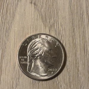 May include: A silver-coloured United States quarter coin featuring the profile of George Washington facing right. The coin is inscribed with the word LIBERTY at the top, the phrase IN GOD WE TRUST on the left, and the year 2023 with a mint mark below.