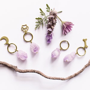 May include: A collection of handmade keychains with raw amethyst crystals. Each keychain features a gold-colored charm in the shape of a moon, paw print, or bone. The keychains are arranged with purple flowers and a twig on a white background.