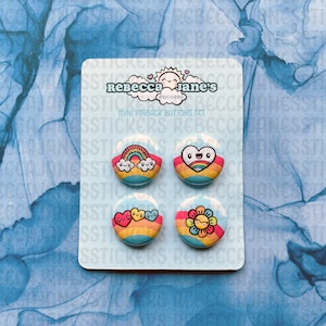 May include: A set of four mini pinback buttons featuring rainbow-themed designs. The buttons are on a blue card with the text "Rebecca Jane's Stickers" and "Mini Pinback Buttons Set".