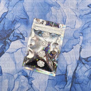 May include: A silver, resealable bag filled with black glitter and star-shaped confetti. The bag features three white cloud-shaped stickers with smiling faces and rain drops. The background is a blue and white abstract design.