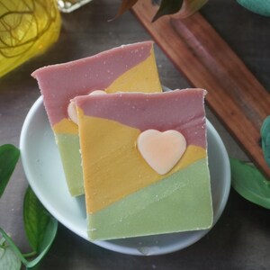 Pode incluir: Duas barras de sabão artesanais com um design em camadas em rosa, amarelo e verde, cada uma encimada por um detalhe em forma de coração. Os sabonetes estão em um prato de cerâmica branco, com vegetação ao fundo.