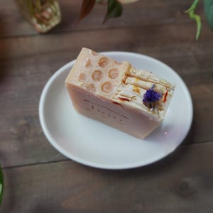 May include: A light pink, rectangular bar of soap with a honeycomb pattern on top, resting on a white plate. The soap is decorated with dried flowers and a small purple flower. The background is a wooden surface.