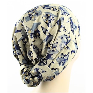 May include: A beige turban with a repeating pattern of blue and gold origami-style shapes. The fabric is gathered and knotted at the front, creating a stylish head covering. The turban is designed to be worn on the head.
