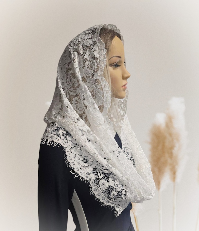 May include: White lace mantilla with a delicate floral pattern. The mantilla is draped over a mannequin's head and shoulders.