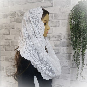 Puede incluir: Mantilla de encaje blanco con borde festoneado y bordado floral. La mantilla está drapeada sobre la cabeza y los hombros de un maniquí.