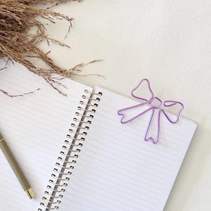 May include: An open spiral-bound notebook with white lined pages, a tan pen with gold accents resting on the left page, and a purple wire paper clip shaped like a bow attached to the top right corner of the right page.