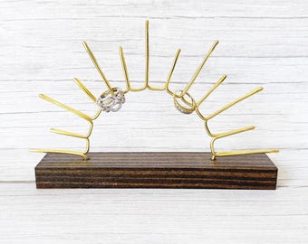 May include: A gold-toned metal jewelry display stand shaped like a sunburst, featuring multiple radiating prongs for holding rings. The metal structure is securely mounted onto a rectangular, dark brown wooden base with visible wood grain texture.