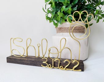 May include: A gold-colored wire sculpture featuring the name Shiloh and the date 3.11.22, topped with a paw print outline. The wire art is mounted into a rectangular, dark-stained wooden base, creating a freestanding decorative display piece.