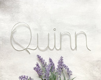 May include: A silver-colored wire sculpture forming the name Quinn in a continuous cursive script, featuring a small circular loop above the letter i, accompanied by a cluster of purple lavender flowers positioned below the wire lettering.