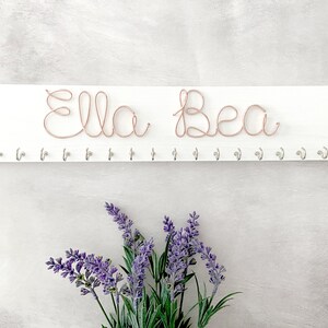 May include: A white wooden wall organiser with the name "Ella Bea" crafted from copper wire. Below the name are several silver hooks. The image includes purple lavender flowers.
