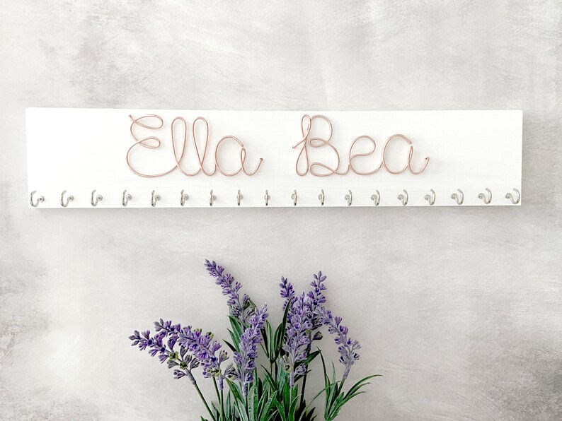 May include: A white wooden wall organiser with the name "Ella Bea" crafted from copper wire. Below the name are several silver hooks. The image includes purple lavender flowers.