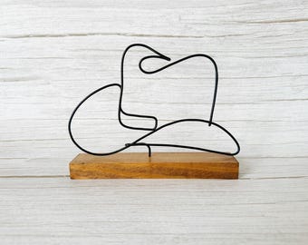 May include: A black metal wire sculpture of a cowboy hat mounted on a rectangular wooden base. The hat's outline is clearly defined against a light wood-grain background. A decorative accent for a rustic or western-themed space.