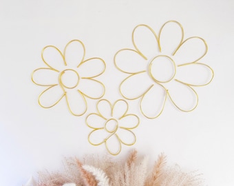 May include: Three gold-colored wire flower shapes of varying sizes arranged in a cluster. Each flower is formed from a single continuous piece of wire, featuring a circular center and several rounded petals radiating outward in a simple outline design.