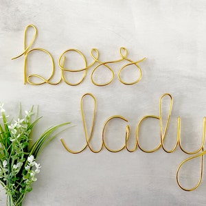 May include: Gold wire sculpture of the words "boss lady" on a white background.