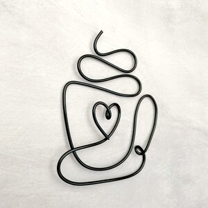 Puede incluir: Escultura de alambre negro de una taza de caf&eacute; con un coraz&oacute;n en su interior. La escultura est&aacute; hecha de una sola pieza de alambre y est&aacute; dise&ntilde;ada para parecer una taza de caf&eacute; con vapor que sale de la parte superior.