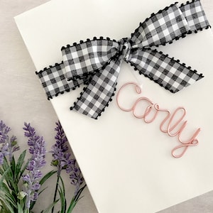 May include: A white gift box with a black and white gingham ribbon tied in a bow. The name "Carly" is written in rose gold wire on a tag attached to the ribbon.