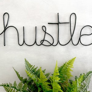May include: Black wire word art spelling out the word "hustle" on a white background. A green fern plant is in the bottom of the image.