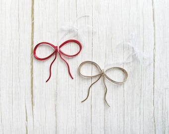 May include: Two wire bows, one red and one gold, rest on a white wooden surface. The red bow has a matching red wire tail, while the gold bow has a gold wire tail. Both bows have a sheer white ribbon attached.