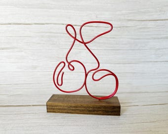 May include: A red wire sculpture mounted on a small, rectangular wooden base. The sculpture has a flowing, abstract design with rounded shapes and lines. The wooden base is a dark brown color.