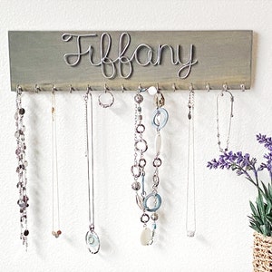 May include: A grey wooden jewellery organiser with the name "Tiffany" in silver wire script. The organiser has hooks for hanging necklaces and other jewellery. Several necklaces of varying styles are displayed.