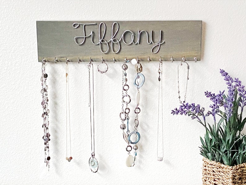 May include: A grey wooden jewellery organiser with the name "Tiffany" in silver wire script. The organiser has hooks for hanging necklaces and other jewellery. Several necklaces of varying styles are displayed.