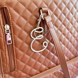 May include: A tan quilted handbag featuring a silver-toned metal letter E charm attached to the strap hardware. The bag includes a vertical zippered pocket with a silver pull, matching tan stitching, and two vertical straps attached to the top.